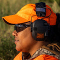 Portrait of a hunter wearing blaze hat and vest, camo jacket, gray safety glasses, and black ear muffs with a black and white american flag patch and an orange logo that reads: TRAIN SMART Everytown Firearms Training.