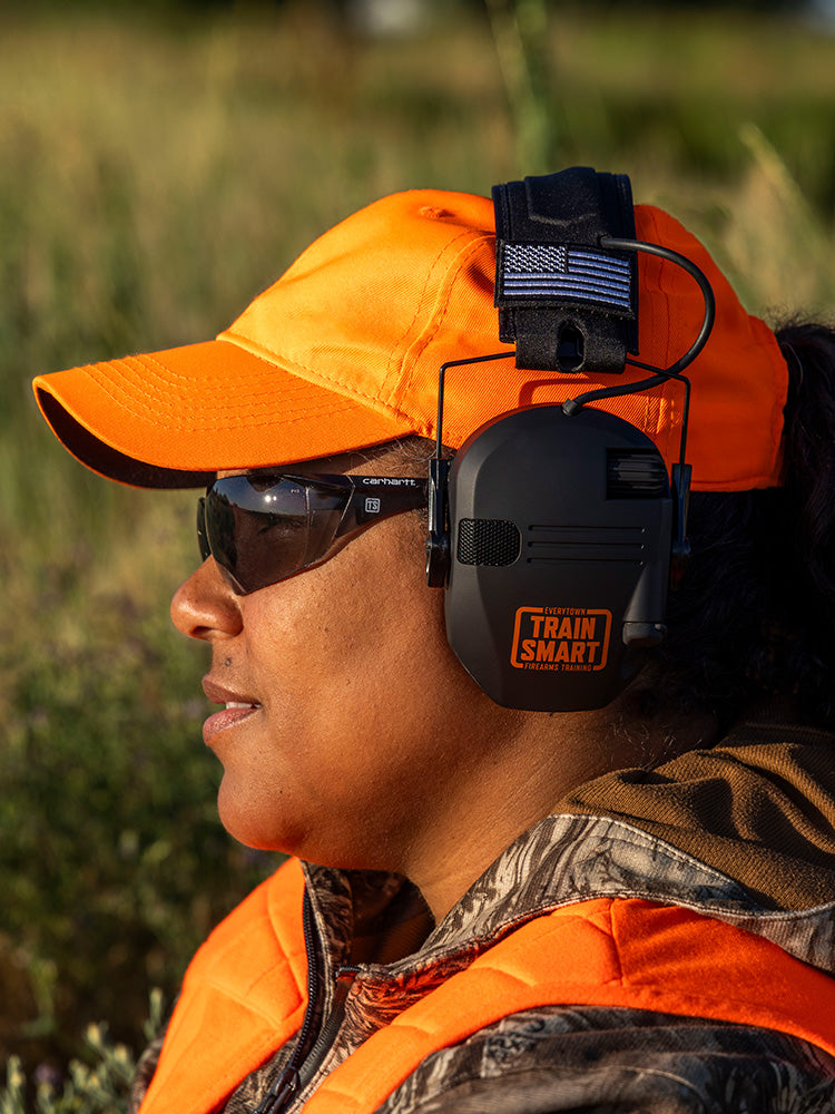 Portrait of a hunter wearing blaze hat and vest, camo jacket, gray safety glasses, and black ear muffs with a black and white american flag patch and an orange logo that reads: TRAIN SMART Everytown Firearms Training.