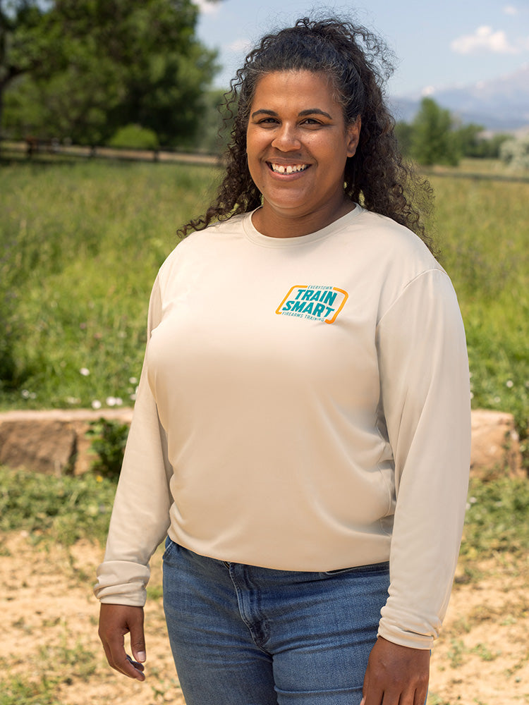 Front view of woman wearing a khaki-colored long sleeve unisex shirt with a teal and orange logo on left chest that reads: TRAIN SMART Everytown Firearms Training.