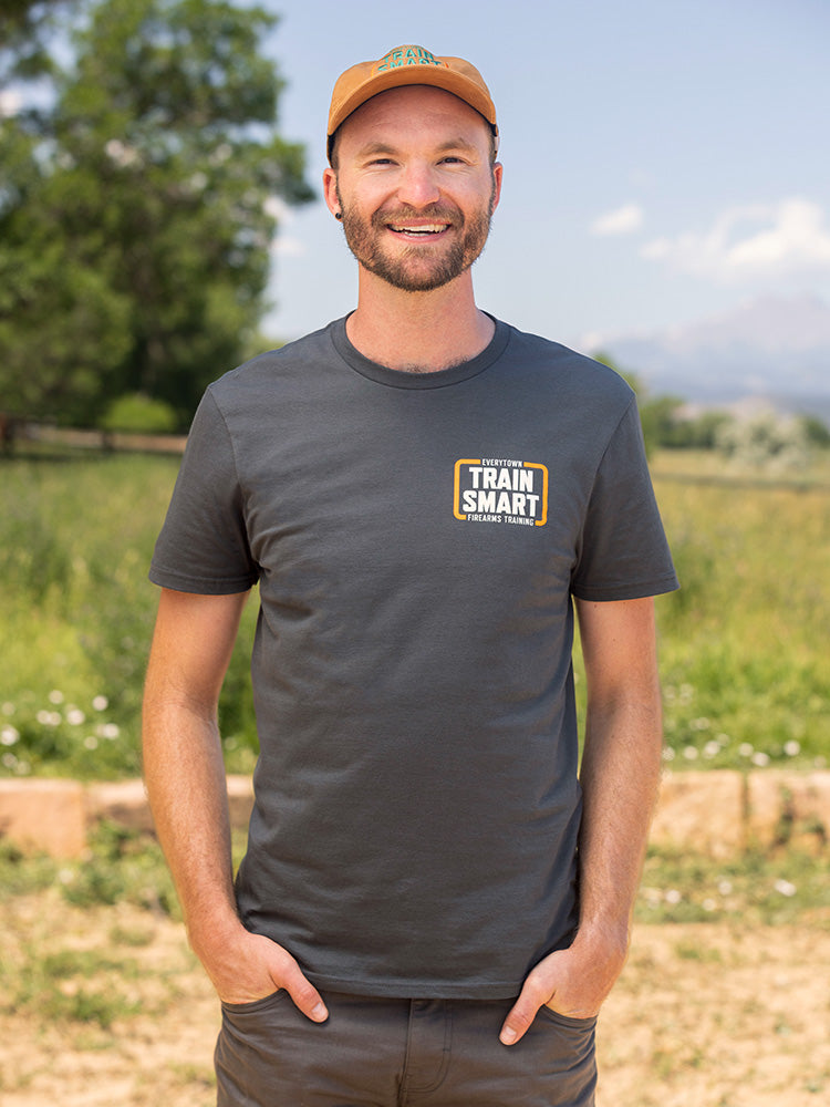 Front view of a man wearing a gray short sleeve unisex t-shirt with a small orange and white logo on left chest that reads: TRAIN SMART Everytown Firearms Training.