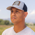 Front view of man wearing a gray trucker hat with black mesh in the back. Embroided orange, white, and black logo on front of hat reads: TRAIN SMART Everytown Firearms Training.