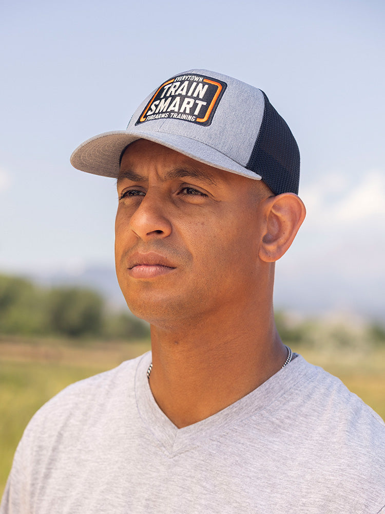 Front view of man wearing a gray trucker hat with black mesh in the back. Embroided orange, white, and black logo on front of hat reads: TRAIN SMART Everytown Firearms Training.